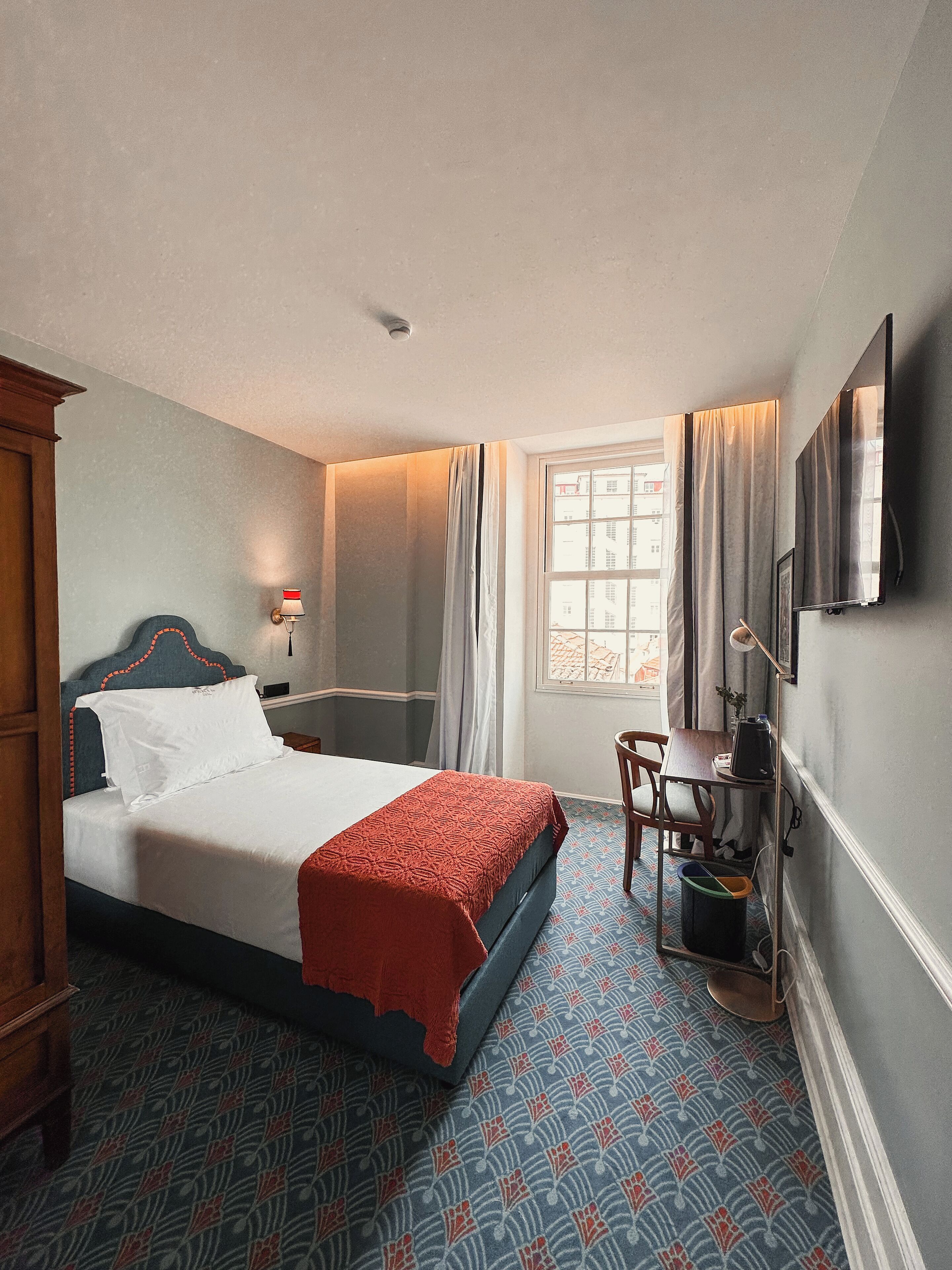 A hotel room with a bed, a desk, a chair, a window with curtains, and a television on the wall.