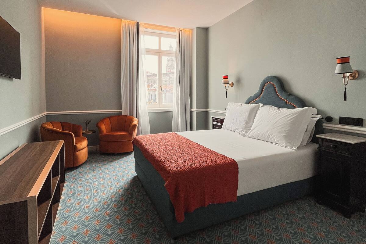 A hotel room with a large bed, a desk, two chairs, a nightstand, and a window with curtains.