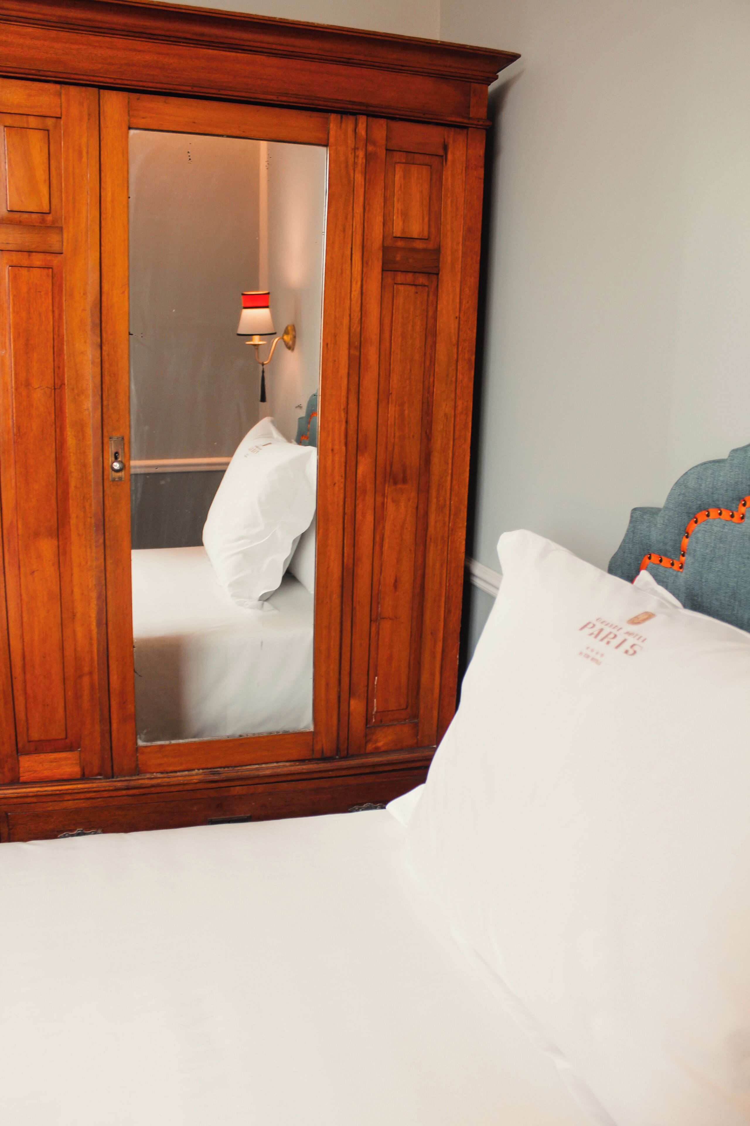 Une armoire en bois avec un miroir, un lit avec une literie blanche et un oreiller bleu avec une étiquette orange.