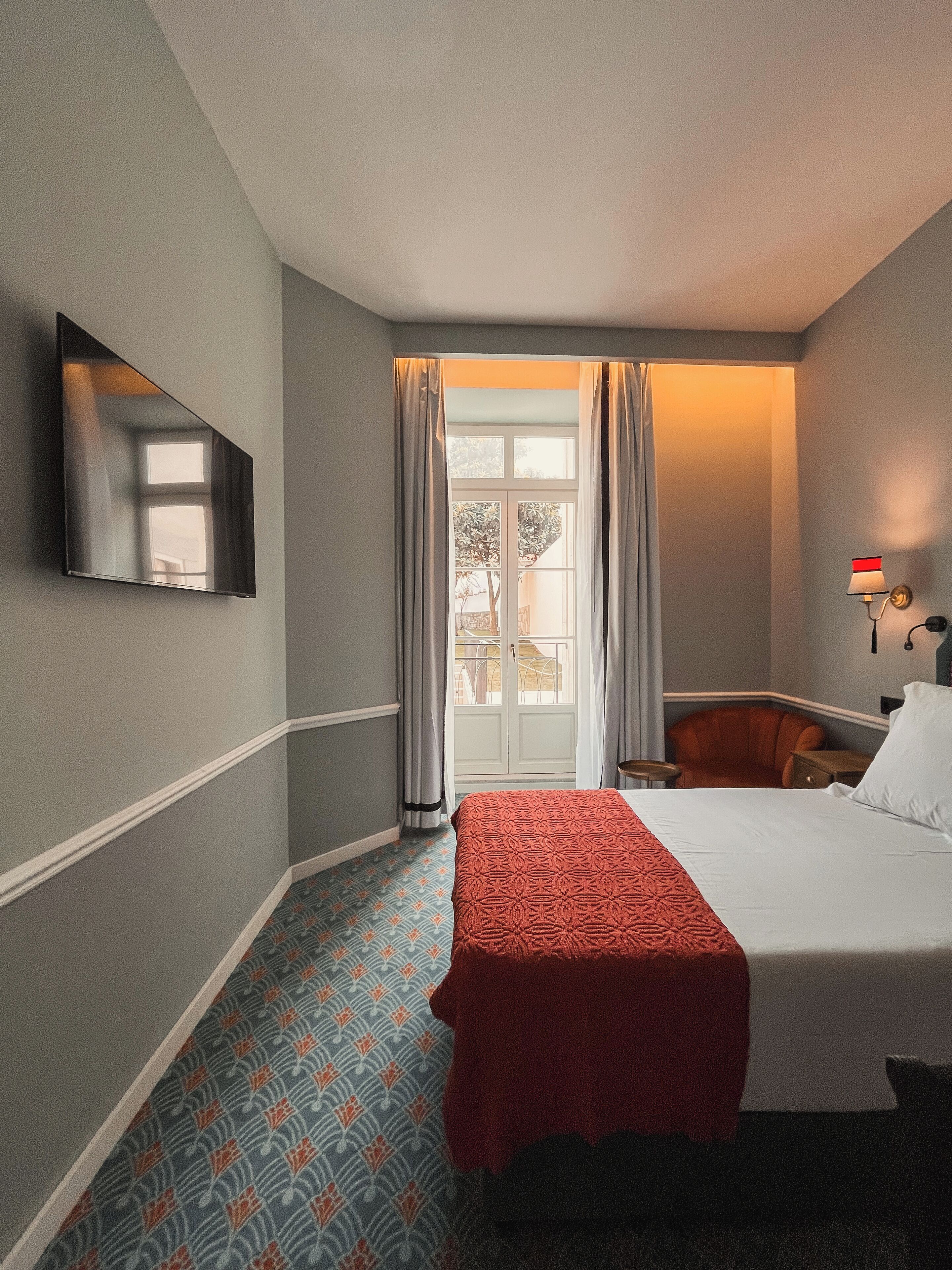 A hotel room with a bed, a chair, a desk, a mirror, and a window with curtains.