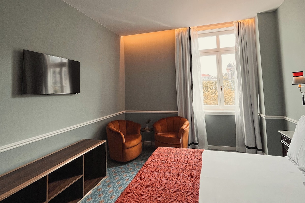 A hotel room with a bed, two armchairs, a TV, a small table, and a window with curtains.