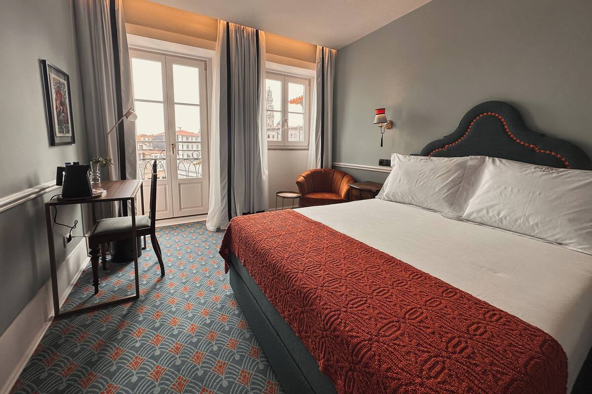 A hotel room with a bed, a desk, a chair, a lamp, and a view of the city through the windows.