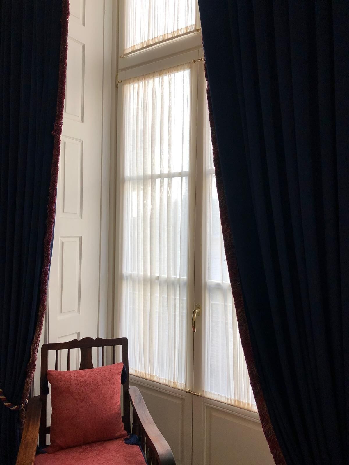 Una habitació amb una cadira de fusta i un coixí vermell, una porta blanca i una finestra amb cortines transparents.