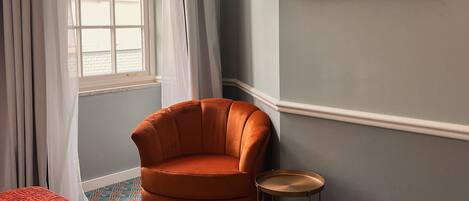 A room with a single orange armchair, a small round table, a flat-screen TV mounted on the wall, and a patterned carpet.