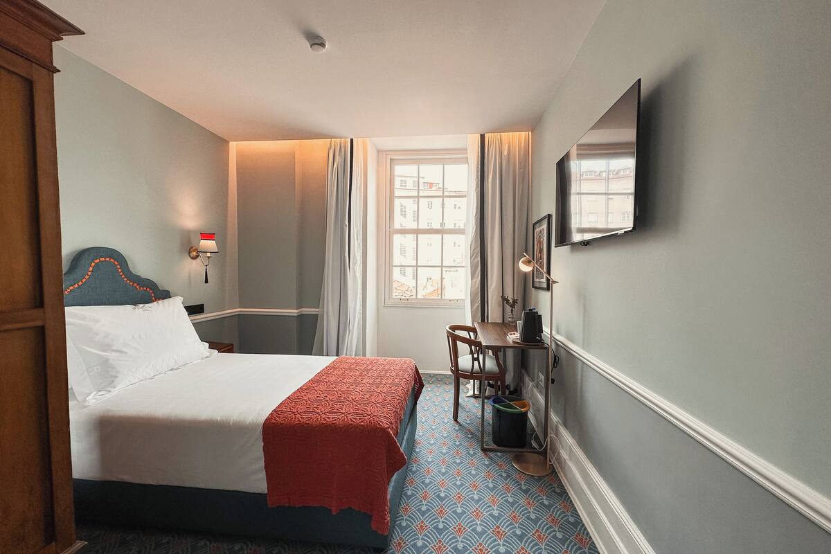 A hotel room with a bed, a desk, a chair, a window with curtains, and a wall-mounted TV.