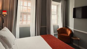 Una habitación de hotel con una cama grande, un plaid rojo estampado, un sillón marrón, una mesita y un televisor instalado en la pared.