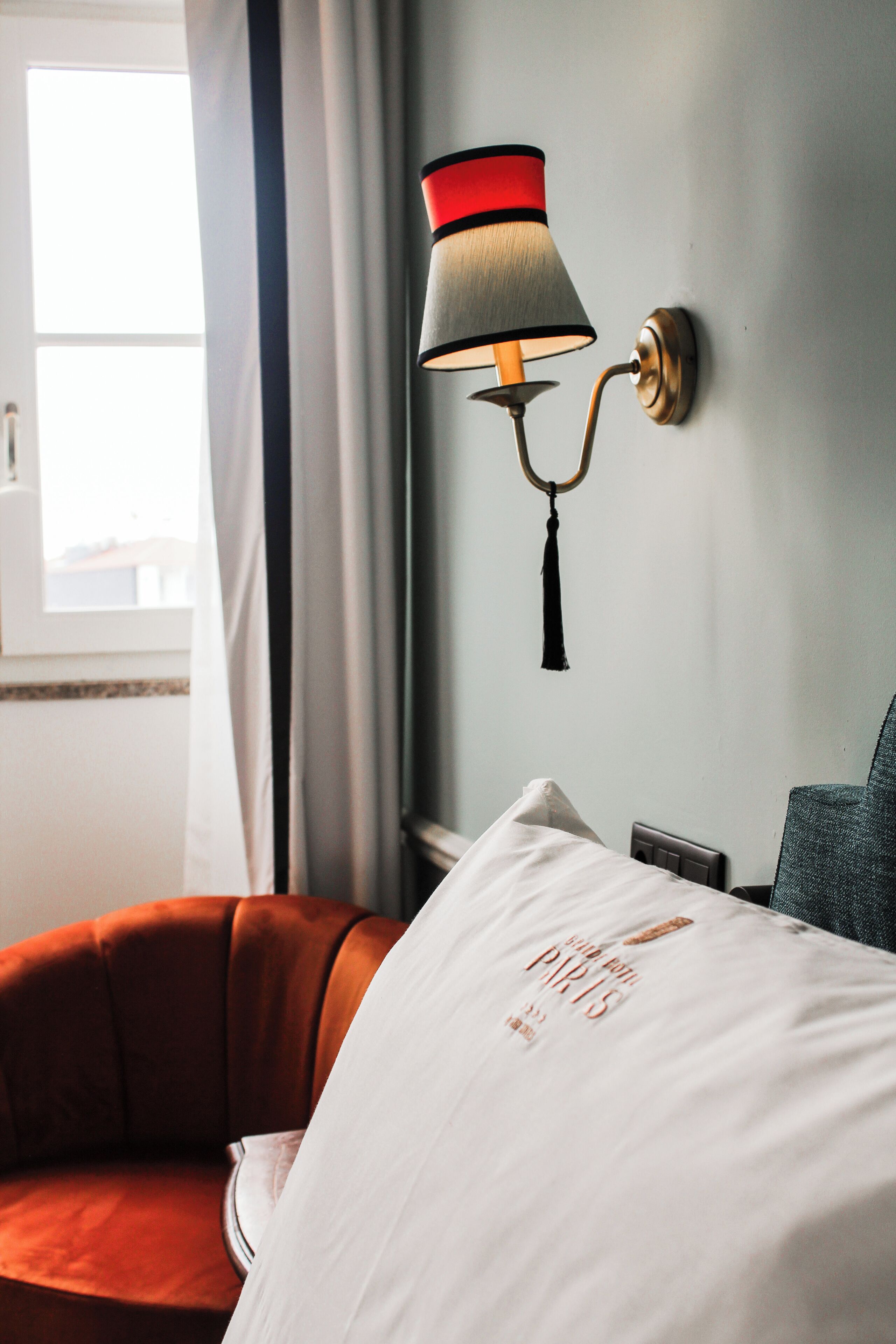 A hotel room with a bed, a wall-mounted lamp, a window with curtains, and a chair.
