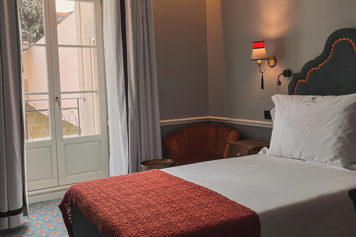 A hotel room with a bed, a chair, a nightstand, a wall-mounted lamp, and a window with curtains.