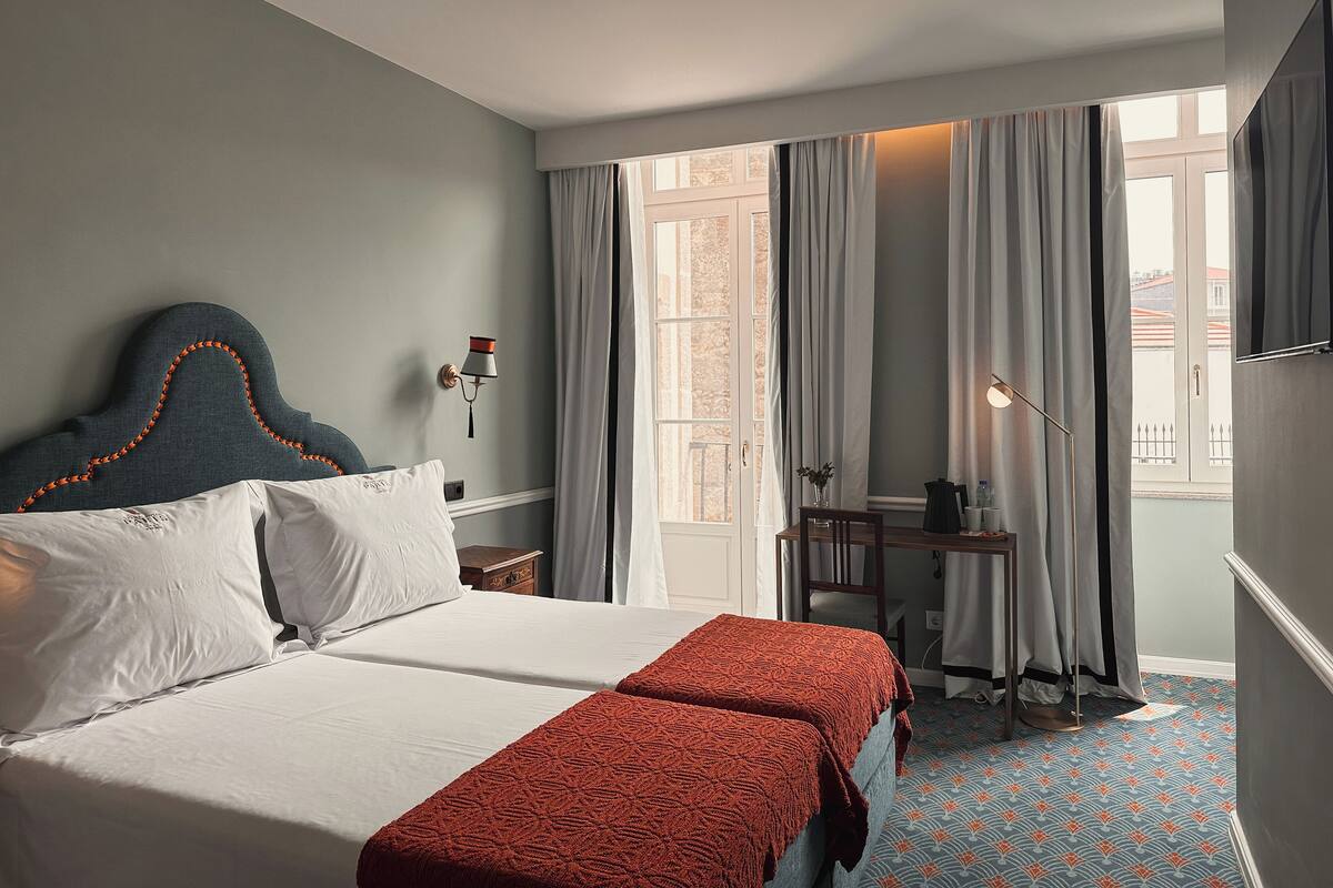 A hotel room with a double bed, a small desk with a chair, a window with curtains, and a patterned carpet.