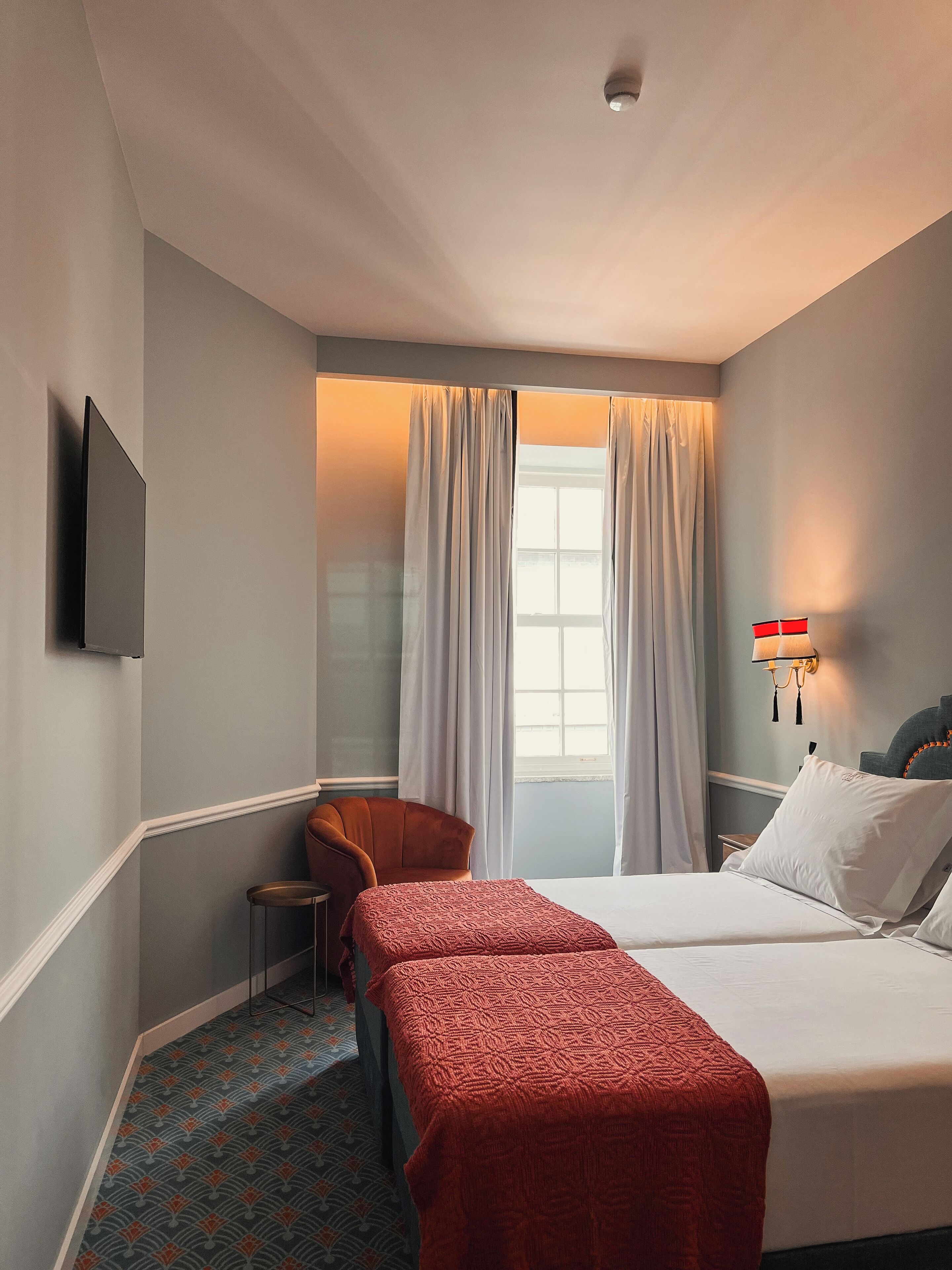 Habitación de hotel con una cama, dos mesitas de noche, una silla, un taburete pequeño, un televisor montado en la pared y una ventana con cortinas.