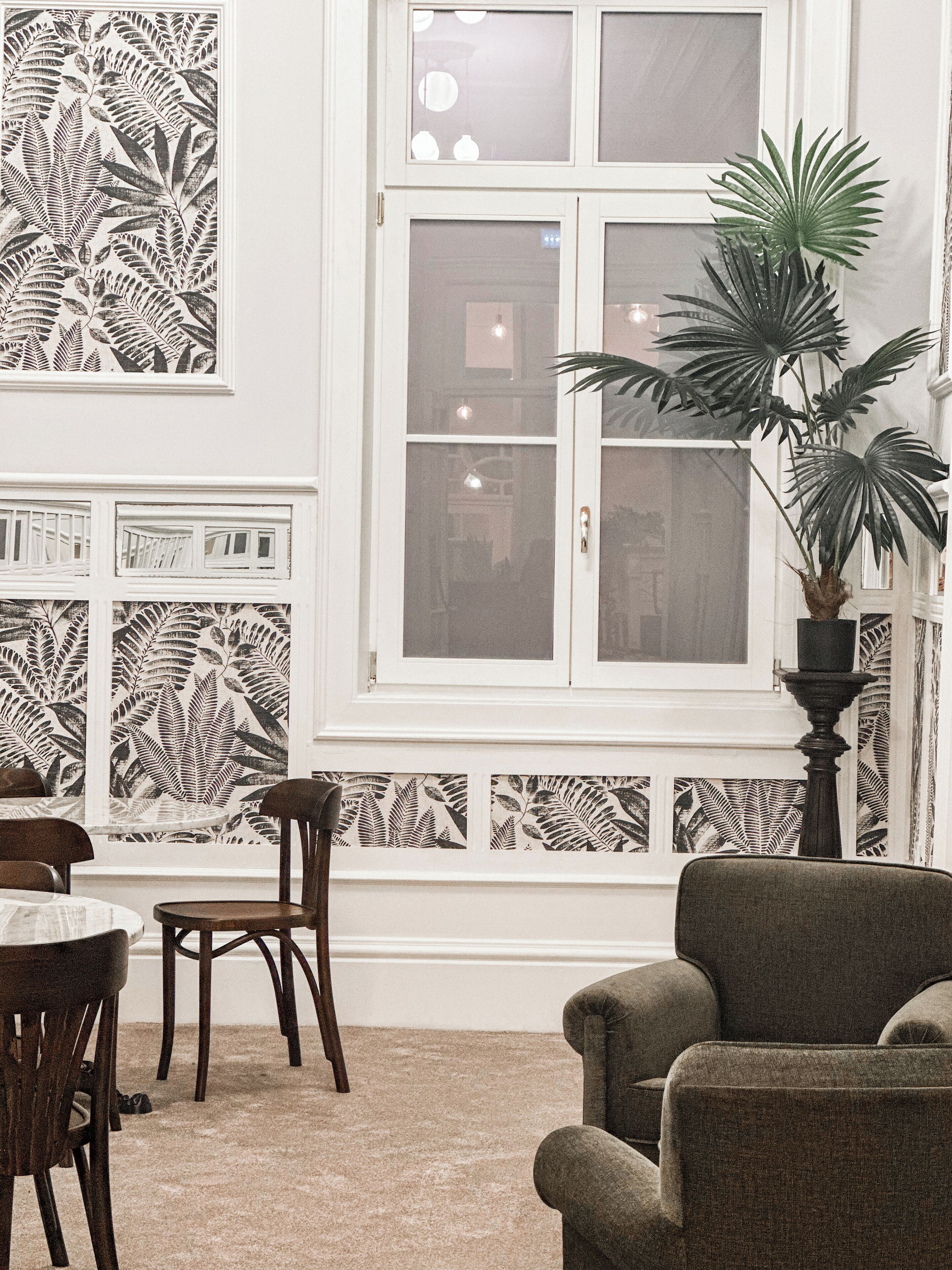 A room with a large window, a potted plant, a dining table with chairs, and a patterned wall.