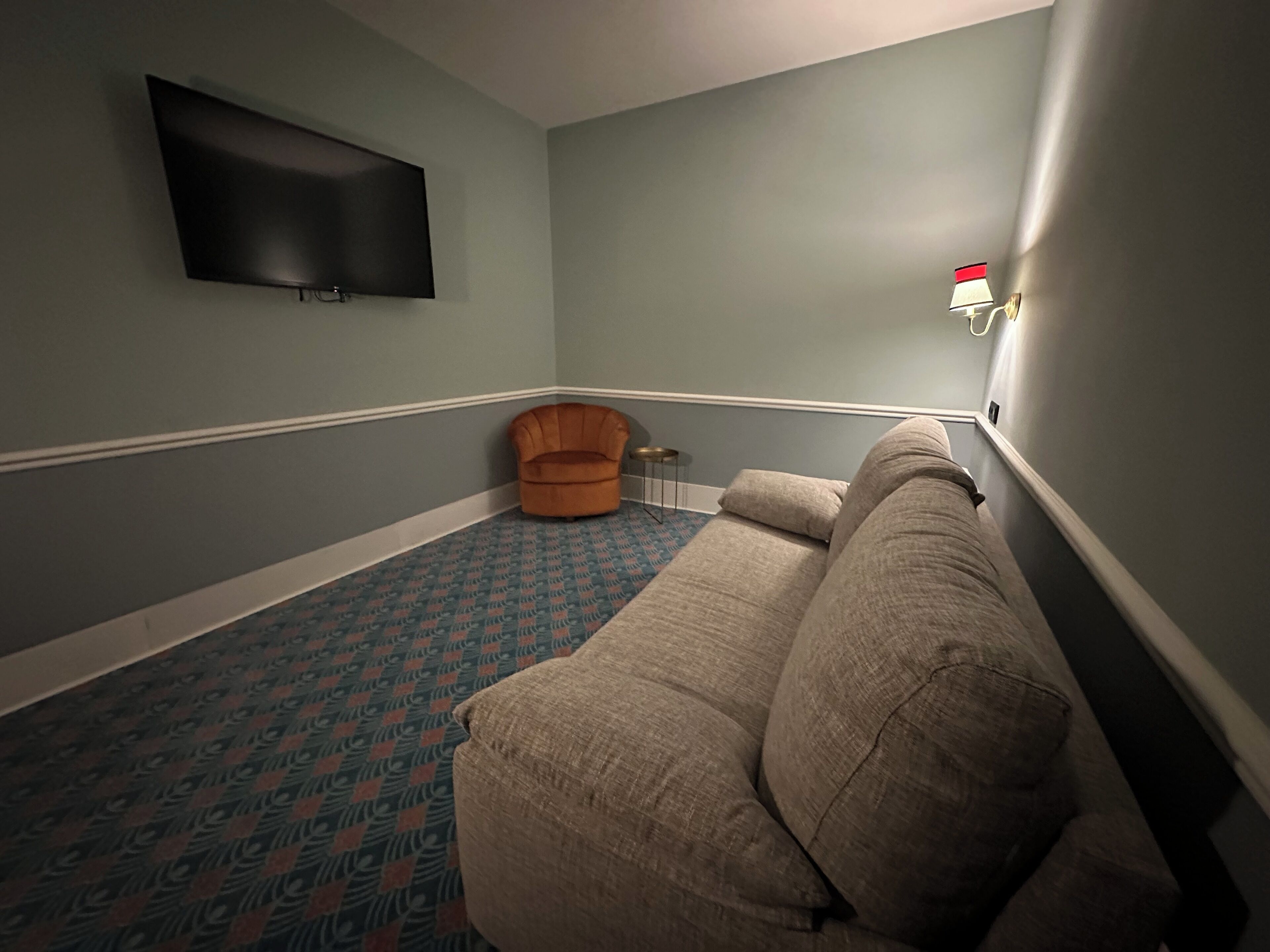 A living room with a patterned carpet, a beige sofa, a small orange chair, and a flat-screen TV mounted on the wall.