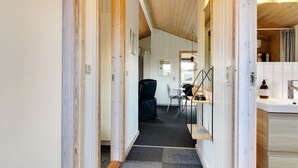 A narrow hallway with wooden walls and a carpeted floor leading to a room with a desk, chair, and a hanging light fixture.