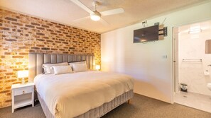A bedroom with a brick wall, a bed with a headboard, a nightstand, a ceiling fan, a TV mounted on the wall, and a bathroom with a toilet and sink.