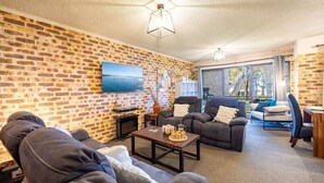 A living room with brick walls, a flat-screen TV, two leather sofas, a coffee table, and a large window with a view of trees. - Bay Parklands, 27/2 Gowrie Ave pool tennis court spa and views (Nelson Bay)
