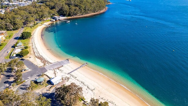 An aerial view of a coastal area with a beach, a pier, and a residential area with houses and a road. - Bay Parklands, 27/2 Gowrie Ave pool tennis court spa and views (Nelson Bay)