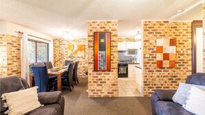 A cozy living room with brick walls, a dining area with a table and chairs, and a kitchen in the background. - Bay Parklands, 27/2 Gowrie Ave pool tennis court spa and views (Nelson Bay)
