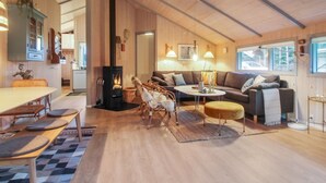 A cozy cabin interior with a central fireplace, a dining area, a sofa, and a window with a view of the outdoors. - "Lovisa" - 500m from the sea by Interhome (Løkken)