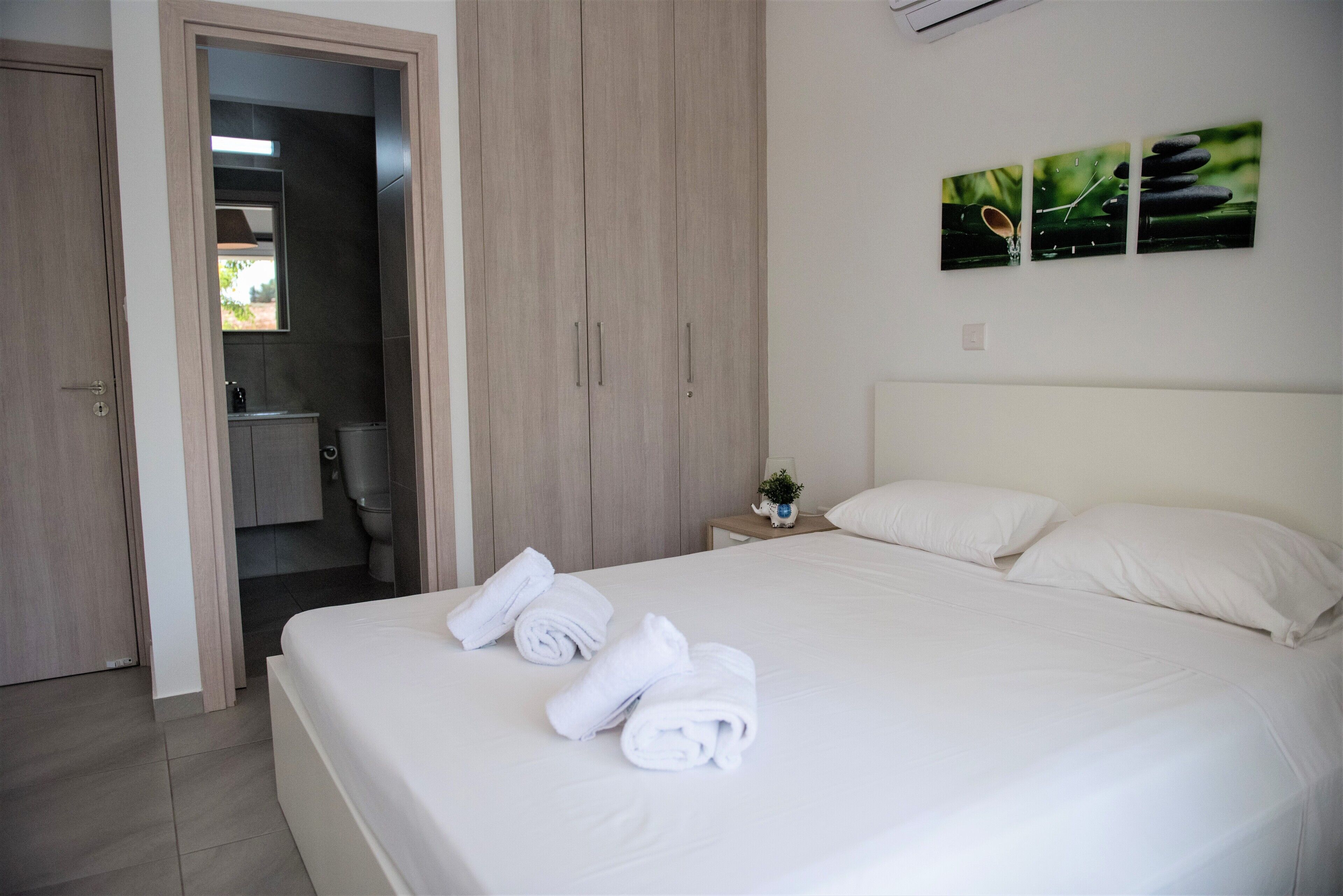 A hotel room with a bed, white towels, a nightstand with a plant, and a wall-mounted artwork.