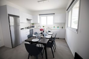A modern kitchen with white cabinets, a stainless steel refrigerator, a microwave, and a dining area with a table set for four. - Carme At The Beach - Mythical Sands Resort (Paralimni)