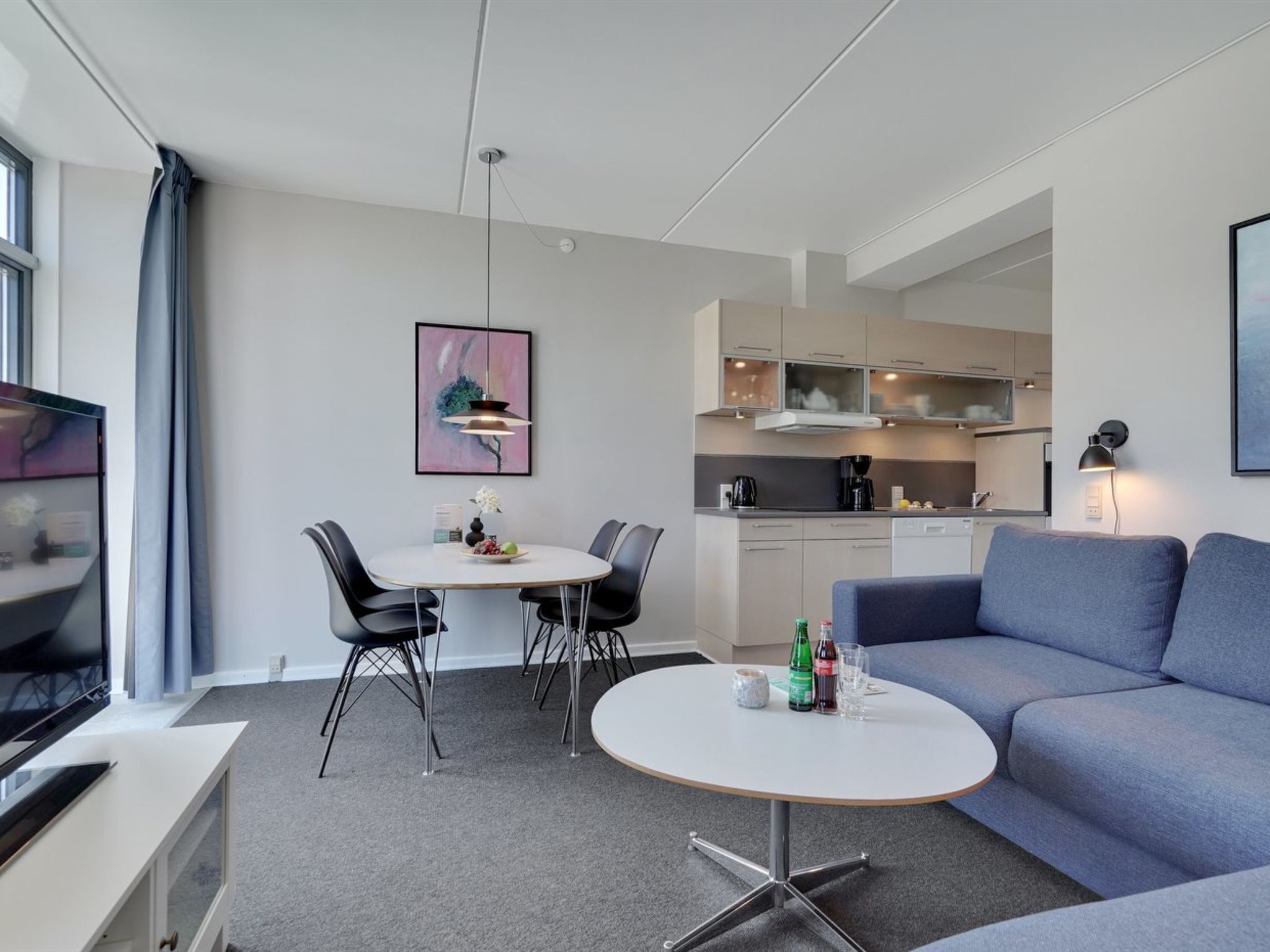 Een moderne woonkamer met een ronde tafel, twee stoelen, een bank, een televisie en een keuken op de achtergrond.