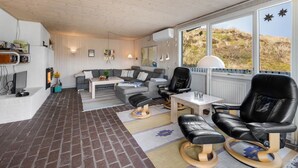 Ein gemütliches Wohnzimmer mit Sofa, Couchtisch und Sesseln, einem Ziegelboden und großen Fenstern mit Blick ins Freie.