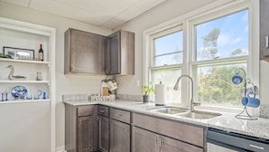 A modern kitchen with dark wood cabinets, a granite countertop, a built-in dishwasher, and a window with a view of trees.