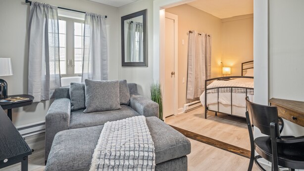 A modern living room with a grey sofa, a matching ottoman, a small table, and a chair. A mirror hangs on the wall, and a door leads to a bedroom with a bed and a nightstand.