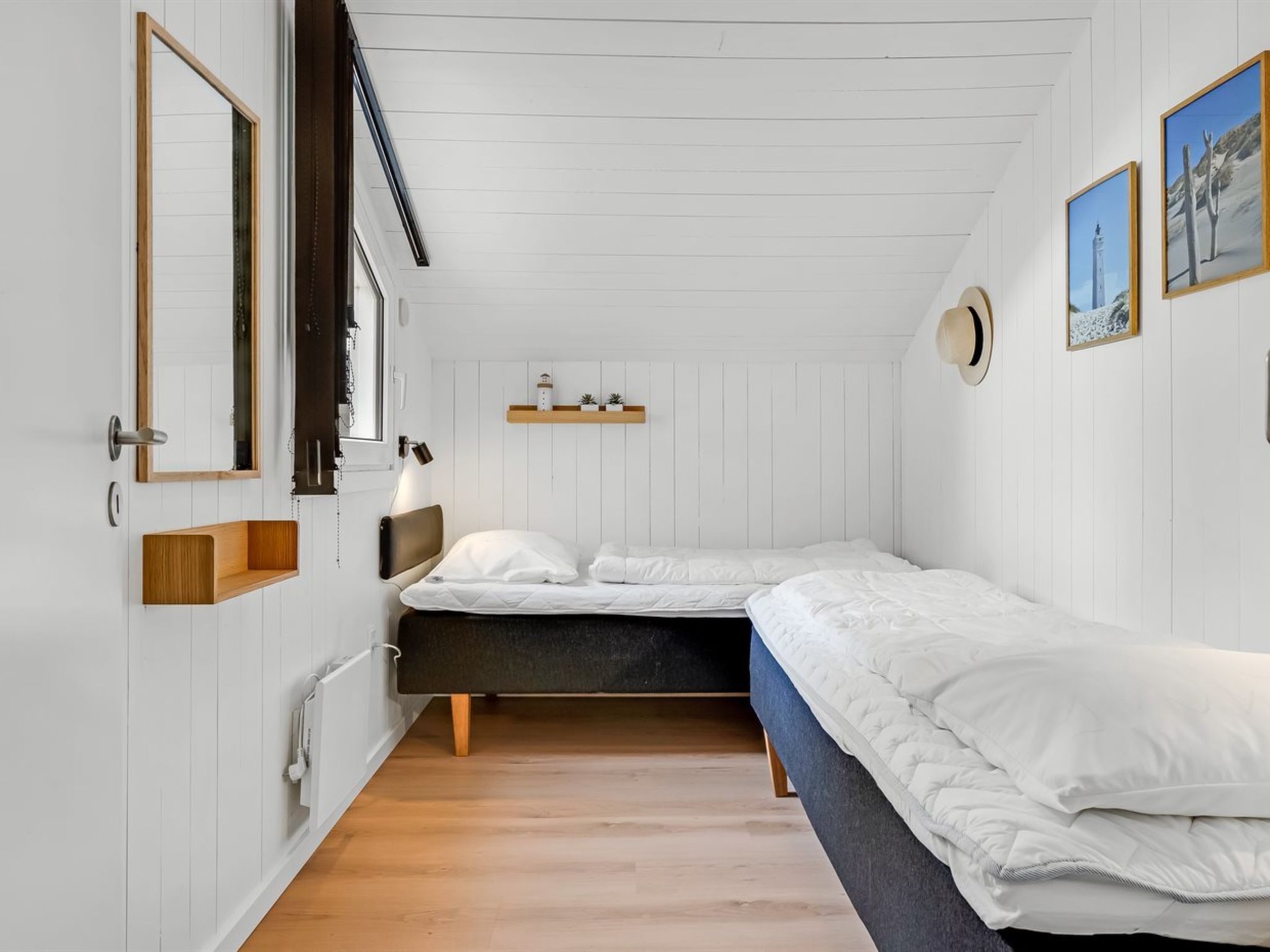 A bedroom with a single bed, a bedside table, a mirror, a shelf with decorative items, and a window with blinds.