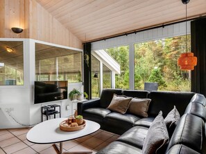 A modern living room with a black leather sofa, a white coffee table, and a large window offering a view of trees.