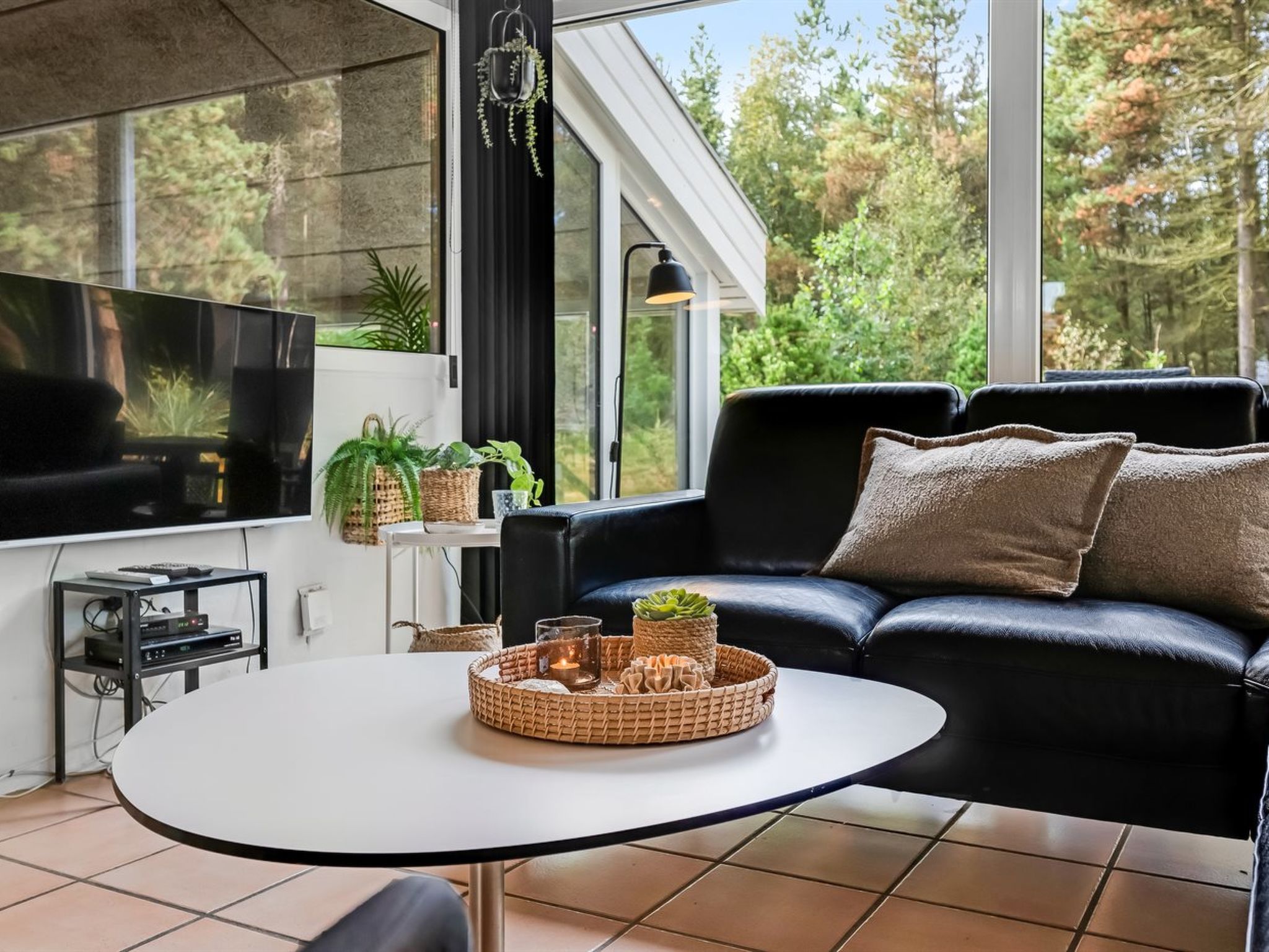 Een moderne woonkamer met een zwarte leren bank, een witte tafel en een televisie aan de linkerkant. Er staan planten en een lamp op de vensterbank.