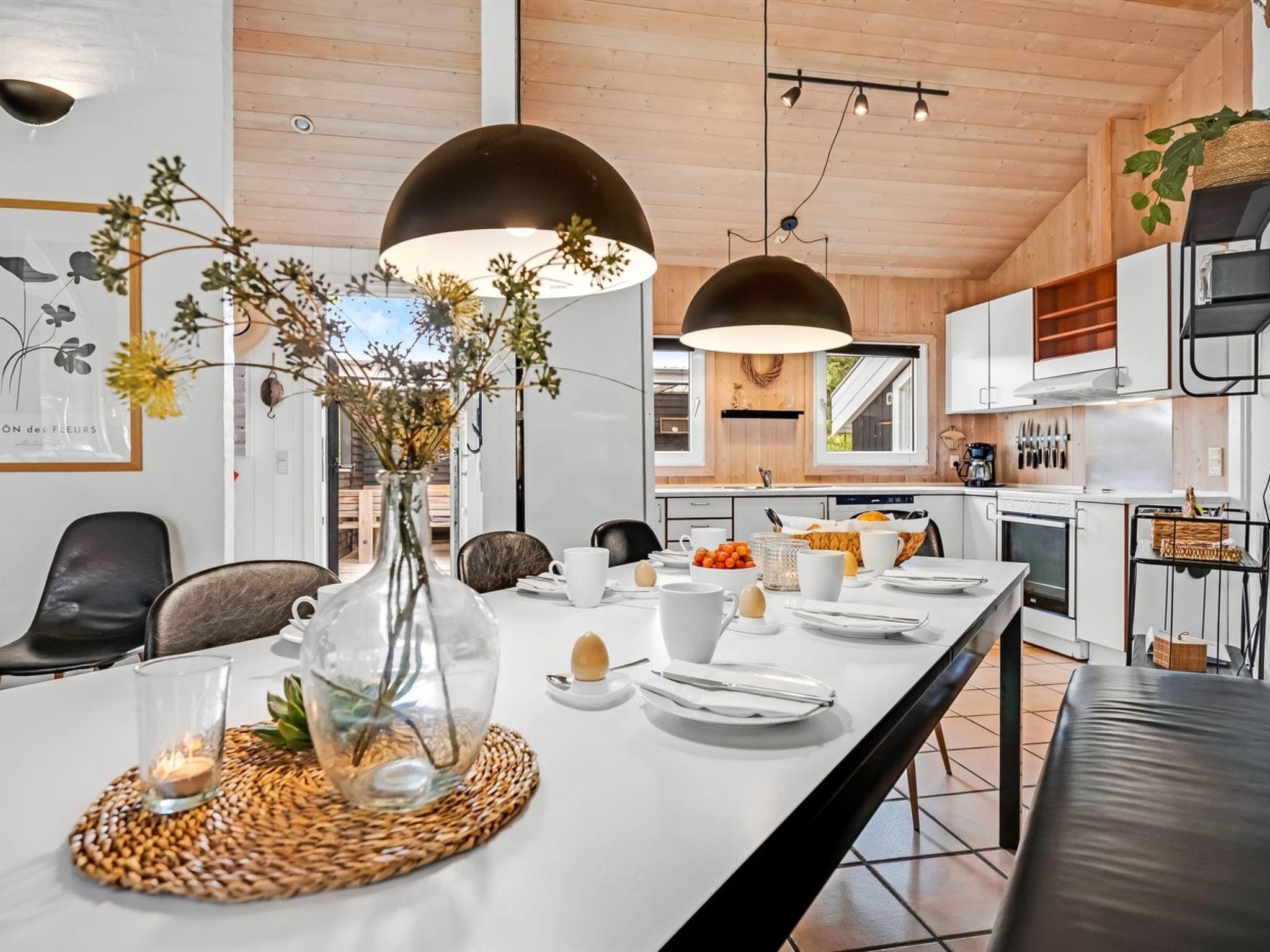 Cozinha moderna com armários brancos, área de jantar com mesa comprida posta para o café da manhã e iluminação pendente.