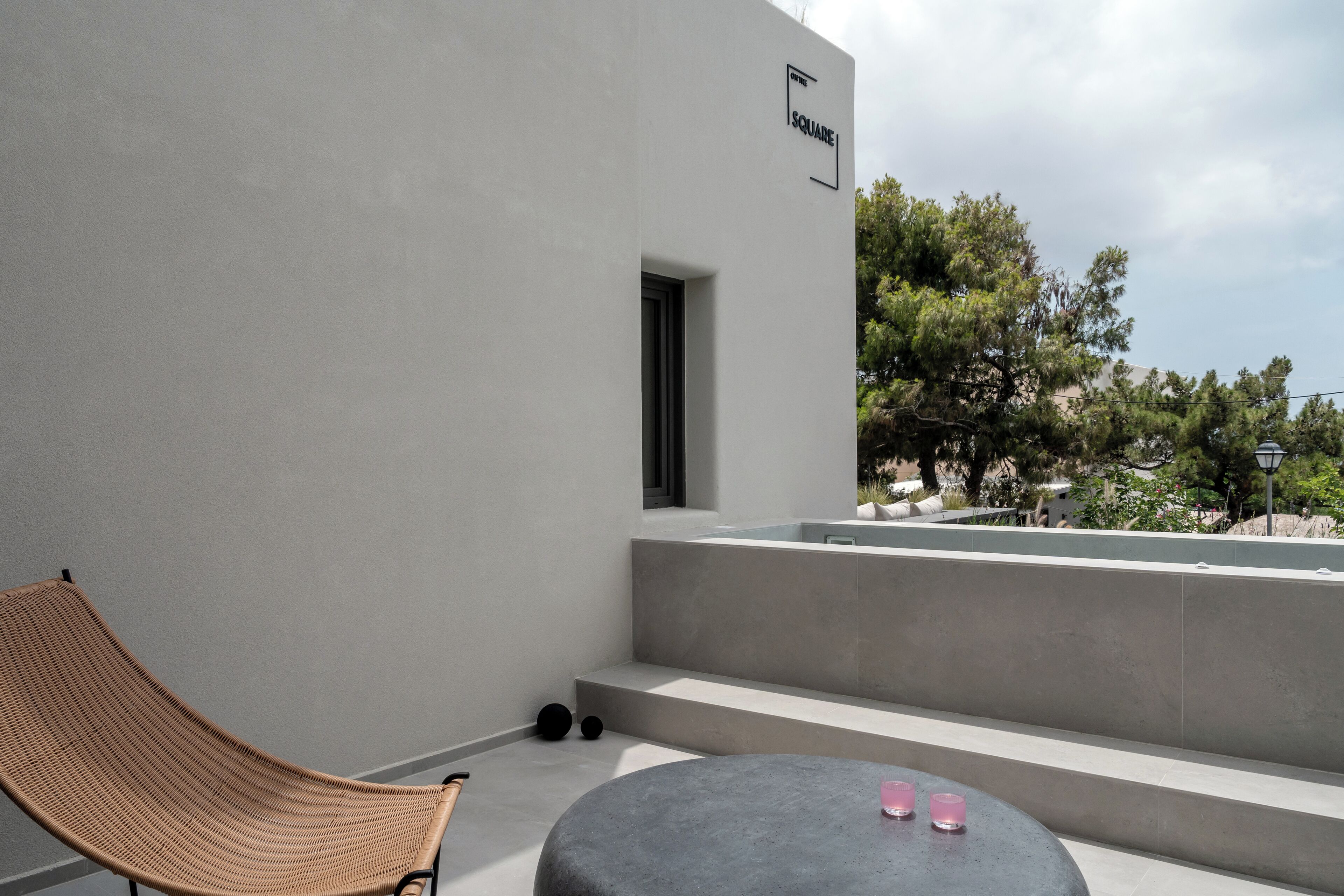 Un espace extérieur moderne avec des sièges, comprenant une chaise en osier et une table ronde, une enseigne indiquant « SQUARE », et une vue sur des arbres et des bâtiments en arrière-plan.