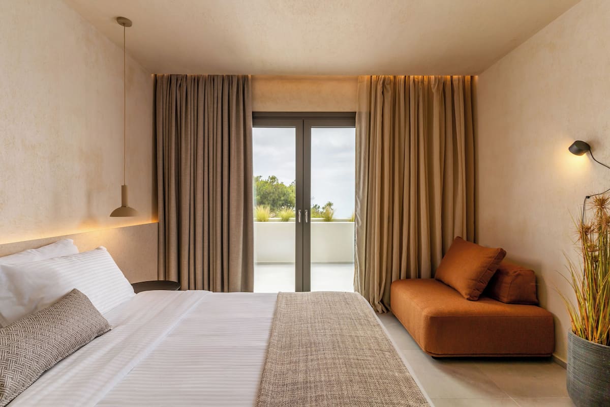 A modern hotel room with a large bed, a sofa, a view through a sliding door, and a potted plant.