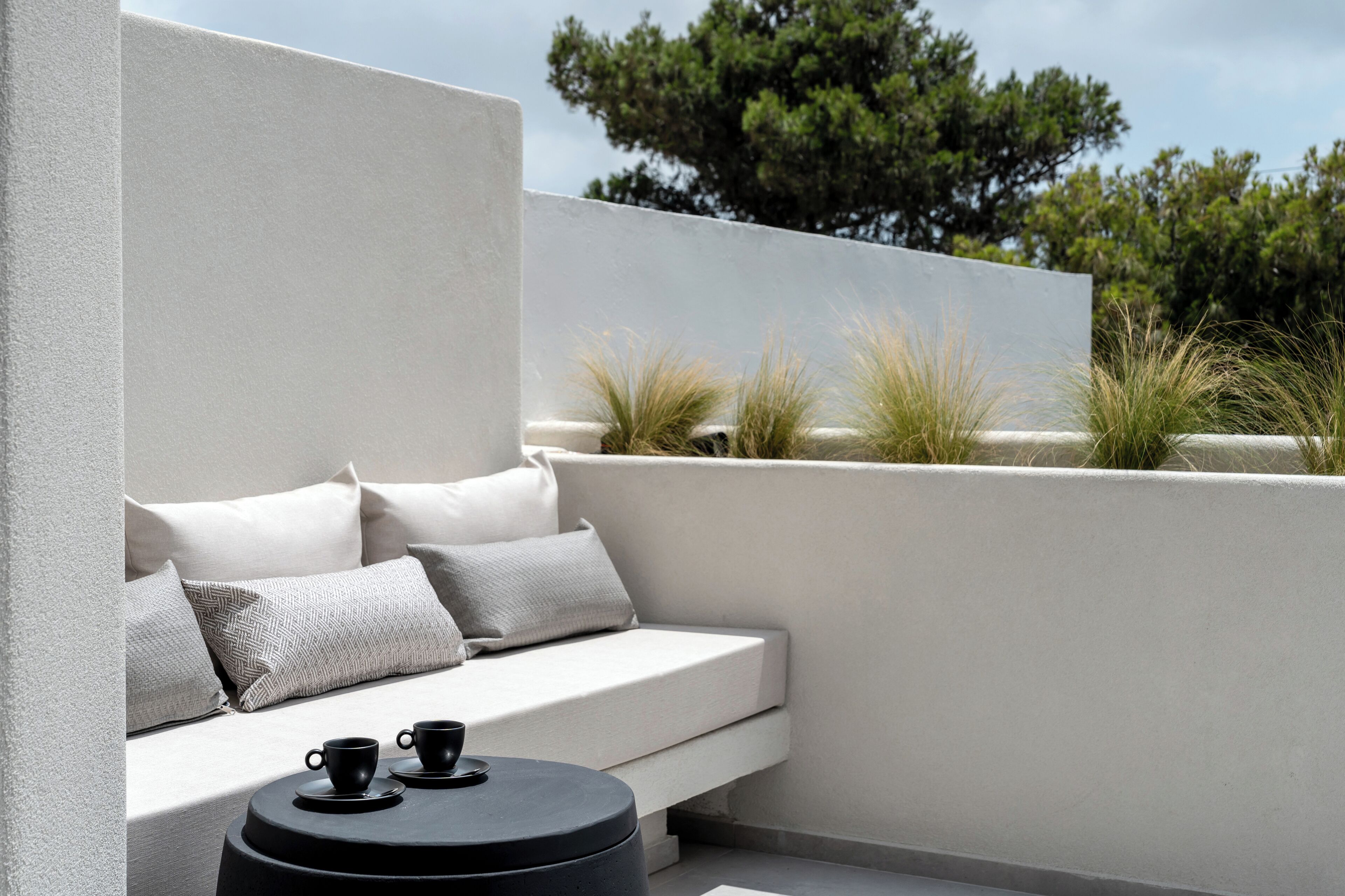 A modern outdoor seating area with a white sofa and cushions, a round black table with two cups, and a view of trees in the background.