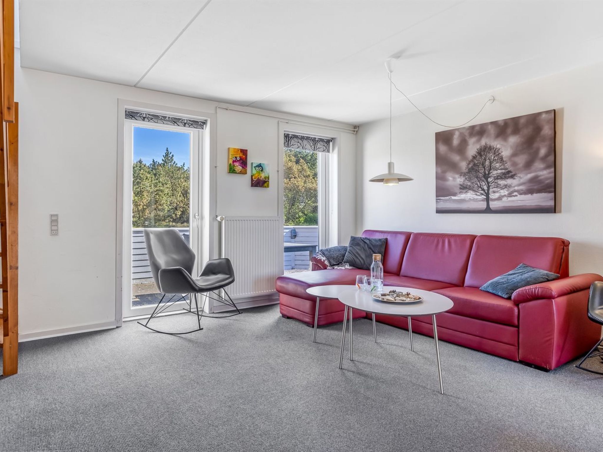 Un salón moderno con un sofá rojo, un sillón mecedora negro y una mesa de centro blanca. Hay un ventanal con vistas a árboles y un techo blanco.