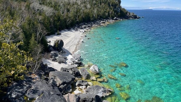 A rocky shoreline with clear turquoise water and a forested hill in the background.