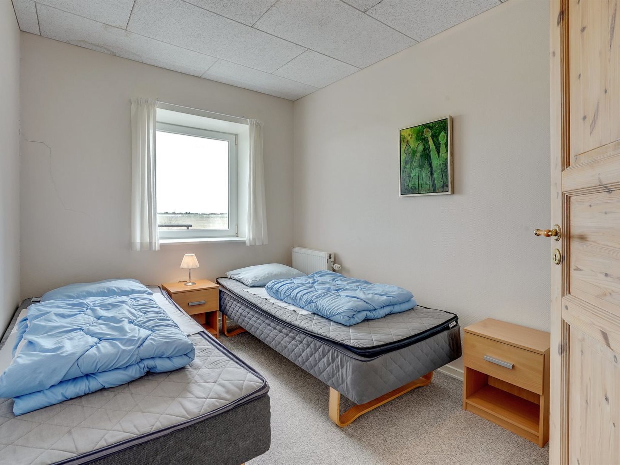 A bedroom with two beds, a window with curtains, a bedside table with a lamp, a small bedside drawer, and a painting on the wall.