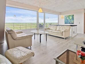 Ett rymligt vardagsrum med ett stort fönster som vetter ut mot en gräsklädd äng, en beige soffa, ett runt soffbord och en tavla på väggen.