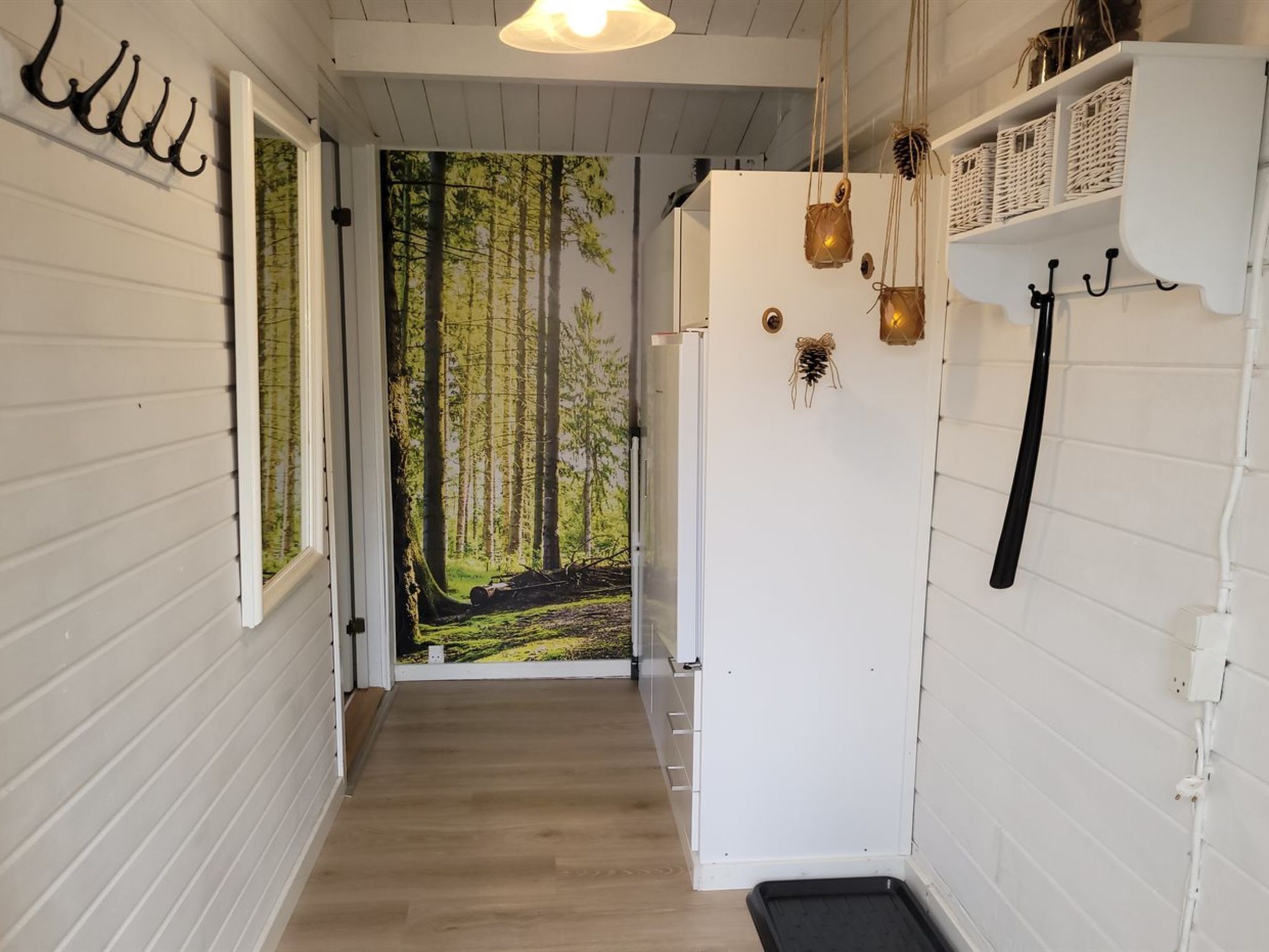 A hallway with a wooden floor, white walls, and a door leading to a forest scene. There are wall-mounted hooks and a shelf with baskets.