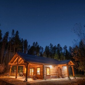 A wooden cabin with a porch, a covered outdoor seating area, and a well-lit entrance.