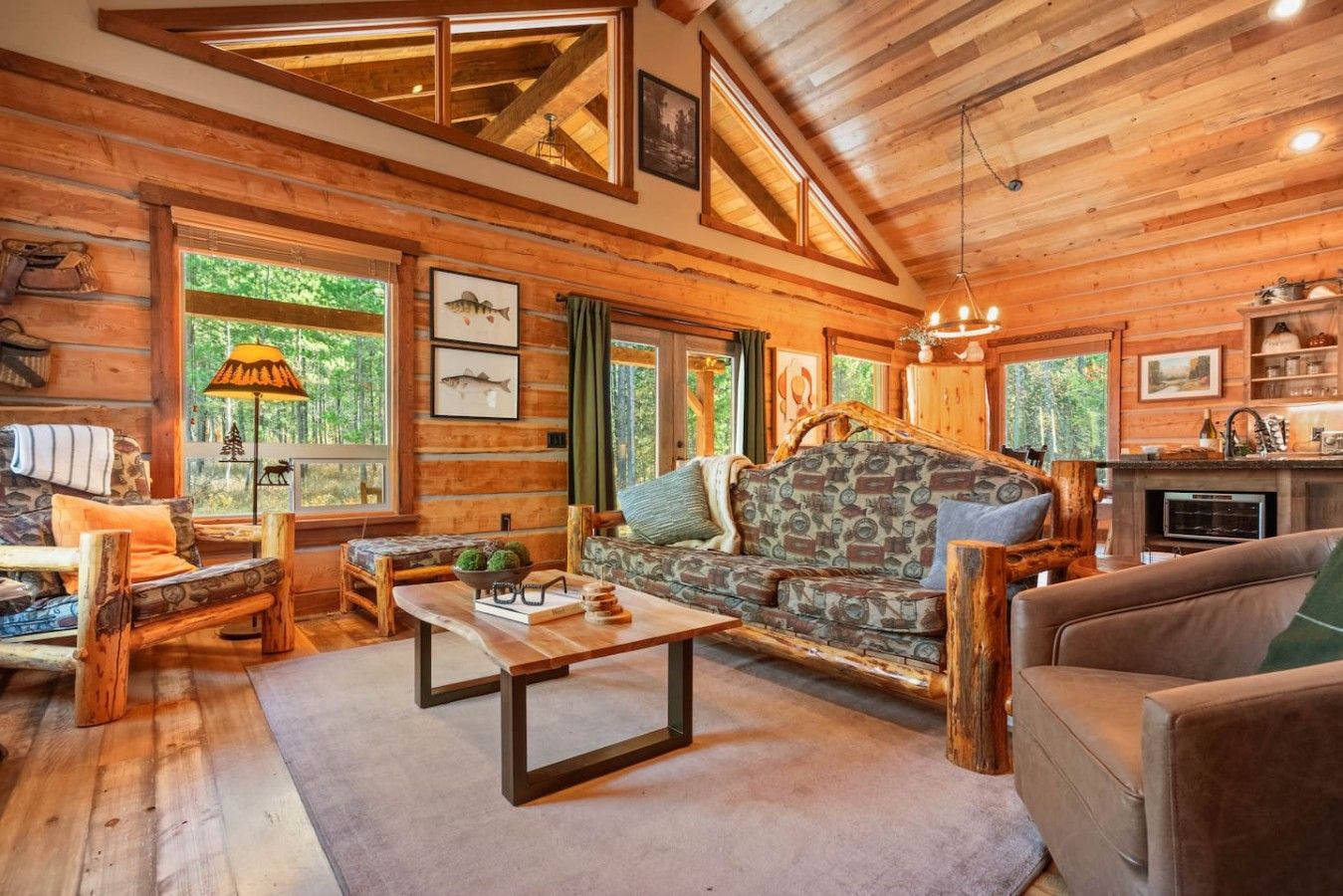 A log cabin interior with a stone fireplace, wooden furniture, and large windows.