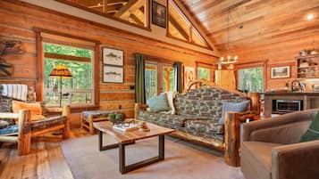 A log cabin interior with a stone fireplace, wooden furniture, and large windows.