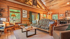 A rustic cabin interior with a wooden ceiling, a stone fireplace, and a cozy seating area with a patterned sofa and a coffee table. - Frontier Cabin, 7 min to Glacier, Fire Pit (Coram)