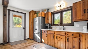 A kitchen with wooden cabinets, a stainless steel refrigerator, and a window with a view of trees. - Saddle n' Sage, 7 min to Glacier, Fire Pit (Coram)