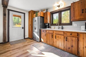 A kitchen with wooden cabinets, a stainless steel refrigerator, a coffee maker, and a window with a view of trees. - Saddle n` Sage, 7 min to Glacier, Fire Pit (Coram)