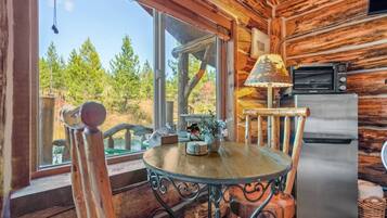 Ein Interieur einer Holzhütte mit einem Esstisch, Stühlen, einem Kühlschrank und einem Fenster mit Blick auf einen Wald.