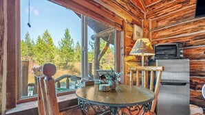 Interior rústico de una cabaña con una mesa de madera, sillas, un refrigerador y una ventana con vista a un bosque.
