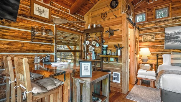 Interior acogedor de una cabaña con paredes de madera, una zona de comedor con taburetes de bar, una cocina con ollas colgantes y una cama con vistas.