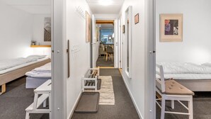 Un pasillo estrecho con dos camas individuales a cada lado, una pequeña zona de estar con una silla y una mesa, y una puerta que conduce a una habitación con escritorio y silla.